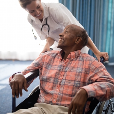 Smiling female doctor looking at disabled senior man sitting on wheelchair against window in retirement home